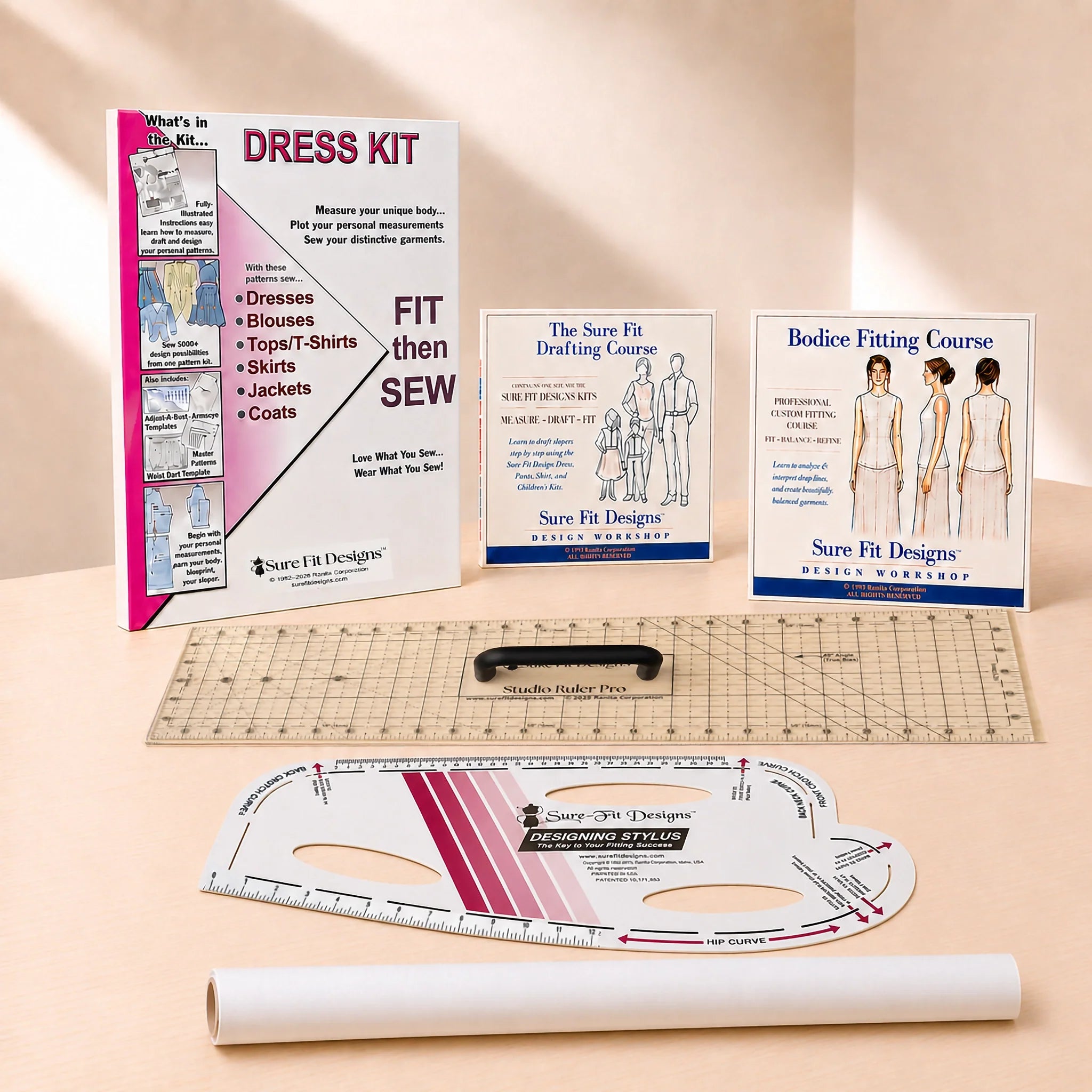A dress-making kit laid out on a light beige surface, smaller course/booklets, a clear ruler, a curved measuring template, and a white fabric roll. The visible brand is Sure Fit Designs, printed on multiple items.