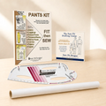 A pants pattern drafting and fitting kit set on a light wood surface with a warm beige background. Includes a roll of tracing paper or vellum, a curved ruler, and two printed booklets: one for 'Pants Kit' and the other 'The Sure Fit Drafting Course'.