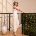 A woman stands on a balcony wearing a white tank top and light beige cargo-style pants with side pockets. She is leaning against a black metal railing next to a green hedge screen and a large beige vase with dried branches.