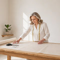 A woman with blonde hair, wearing a white blouse and a gold measuring tape necklace, is seen from the waist up as she works at a wooden table, smoothing out a large roll of beige fabric. A pair of scissors and a few pins lie on the table next to the roll.